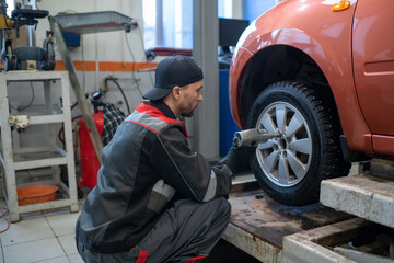 Wall Mural - Young repairman in workwear using electric handtool while sitting on squats and changing wheel of car in garage