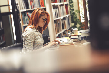 Wall Mural - Red hair female student studying  int the college library. Using laptop.
