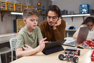 Wall Mural - Boy using digital tablet while sitting at the table with teacher he controlling the robot with his computer at robotics lesson