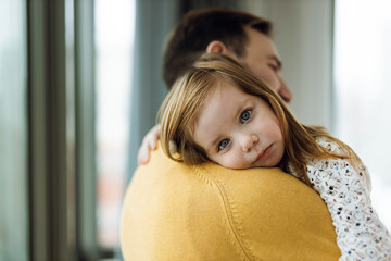 Cute little girl embracing her father by the window. She is looking at camera