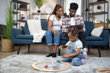 Wall Mural - African american married couple sitting on couch and using laptop while her cute daughter playing on floor. Beautiful family at home. Leisure activity.