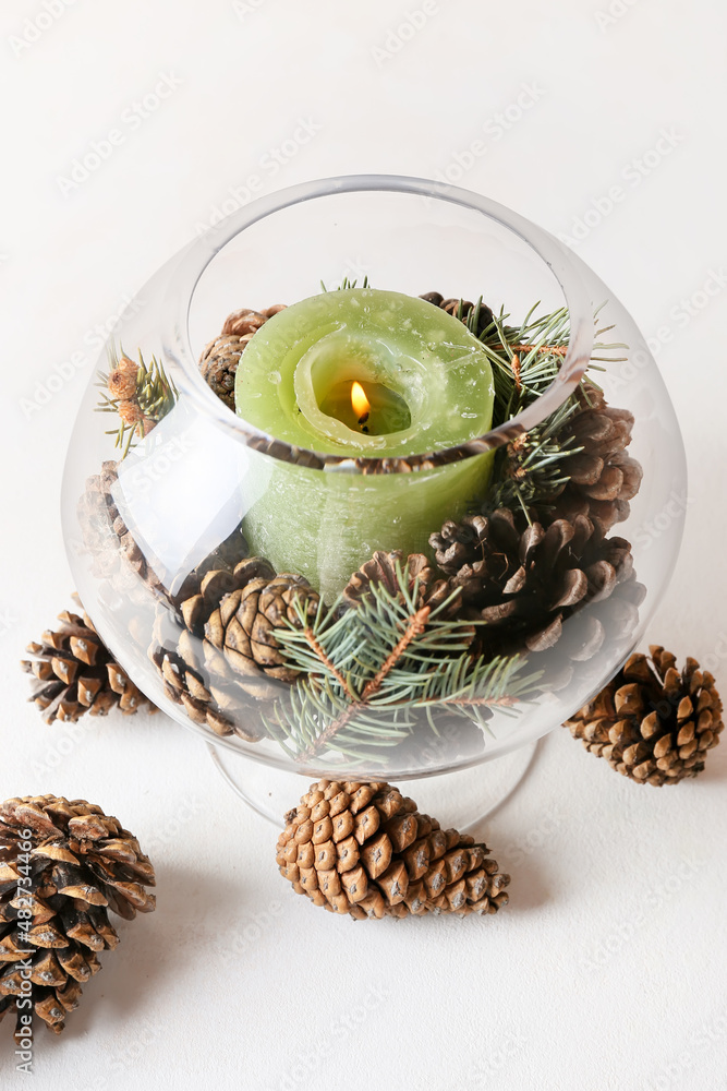 Glass holder with burning candle and pine cones on light background