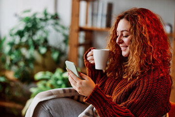 A cute ginger girl with curly hair is sitting in the chair at home in the morning, texting messages on the phone and drinking her coffee.