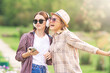 Summer lifestyle portrait of two best girls friends laughing and talking outdoors in park. Wearing casual clothing, sunglasses, headphones and holding smartphone. Enjoying time together.