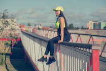 A Woman In Sportswear And A Cap Sits On The Railing Of A Bridge