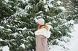 © Dina - Happy woman in winter forest during snowfall, enjoying winter. Millennial young woman with long hair in beige warm outfit walking in the snowy park in nature near fir trees, being careless and free..