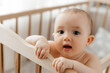 © Ananass - portrait of a 5 month old baby girl standing in a crib in a bright nursery after sleep and looking and smiling at the camera