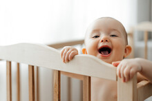 Portrait Of A 5 Month Old Baby Girl Standing In A Crib In A Bright Nursery After Sleep And Looking And Smiling At The Camera
