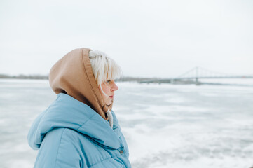 Portrait of a cute girl with blond hair in warm clothes on a background of frozen river and bridge. Winter walk.