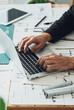© Tijana - High angle view of an anonymous architect sitting at his desk and working with blueprints, sketches, digital tablet while typing new project on his laptop computer