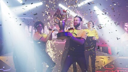 Wall Mural - Bearded leader of cybersport team kissing golden trophy and dancing with team under confetti while celebrating victory in esports gaming championship