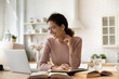 © fizkes - Pretty Hispanic smiling young woman in glasses sit at table in kitchen e-learning using computer, hold pencil take notes, use heap of textbooks. Modern tech, sel-education, video call, tuition concept