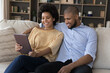 © fizkes - Beautiful African couple relaxing on sofa using digital tablet, choosing online goods, buying e-services at home, booking hotel planning vacation. Modern tech, distancing services, app usage concept