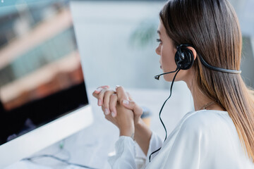 young businesswoman in headset sitting with clenched hands and looking at monitor.