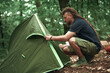 Young hippie hiker setting up his tent in the forest, pitching in a forest clearing, getting ready for a camping sleepover