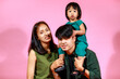 © Bangkok Click Studio - Portrait closeup studio shot of young happy Asian family husband wife and daughter smiling look at camera together on pink background. Kindergarten preschooler baby girl piggyback riding on dad neck