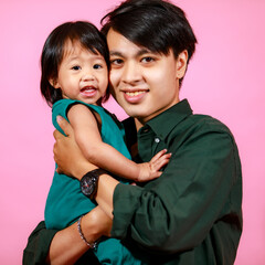 Wall Mural - Portrait closeup studio shot of Asian young happy teenager father holding hugging carrying little cute preschooler kindergarten baby girl daughter smiling look at camera together on pink background