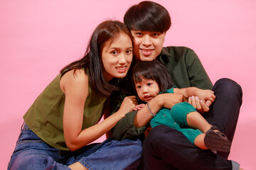 Wall Mural - Portrait closeup studio shot of young happy Asian family father mother and little cute baby girl daughter sitting on floor together smiling look at camera on pink background