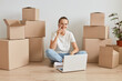 © sementsova321 - Positive woman wearing white T-shirt sitting on the floor near cardboard boxes with personal stuff, talking phone, looking at camera, expressing positive emotions.