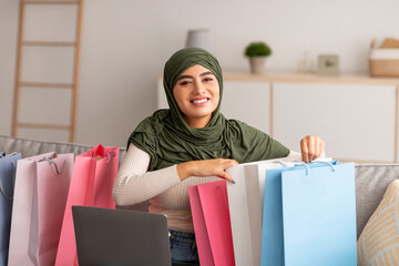 Wall Mural - Happy Arab woman in hijab sitting on couch with laptop and bright shopper bags, buying goods online at home