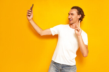 smiling man takes selfie on phone showing V sign of peace victory with hand on camera dressed in casual clothes. isolated on studio yellow background. concept - people, technology, video communication