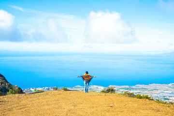 Tourist and freedom lifestyle people travel concept. Woman viewed from back opening arms to enjoy an amazing view and landscape. Blue ocean and sky from a top of a cliff. Happy lady