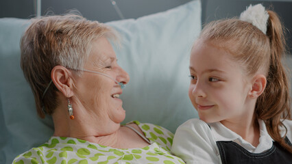 close up of senior woman with illness enjoying visit from litte girl in hospital ward bed. aged pati