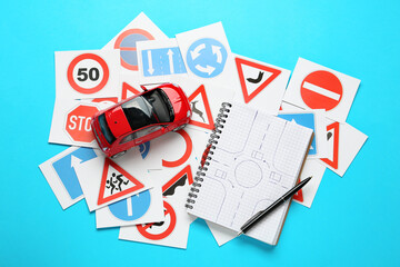 Many different road sign cards, notebook with sketch of roundabout and toy car on light blue background, flat lay. Driving school