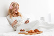 © Prostock-studio - Smiling millennial pretty european blonde woman in robe and headphones reading news, chatting, watch video