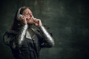 Wall Mural - Young medieval warrior or knight with dirty wounded face in headphones listening to music isolated over dark vintage background. Comparison of eras, history