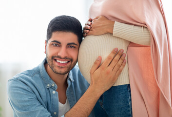 Wall Mural - Caring muslim man putting his ear on wife's pregnant tummy, listening to baby's heartbeat, closeup