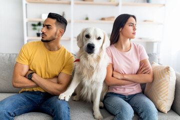 Canvas Print - Upset young multinational couple having fight, looking in opposite directions, cute dog sitting on couch between them