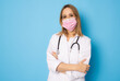 © Danko - Female nurse standing arms crossed. Confident young woman doctor wearing surgical mask isolated on blue background.