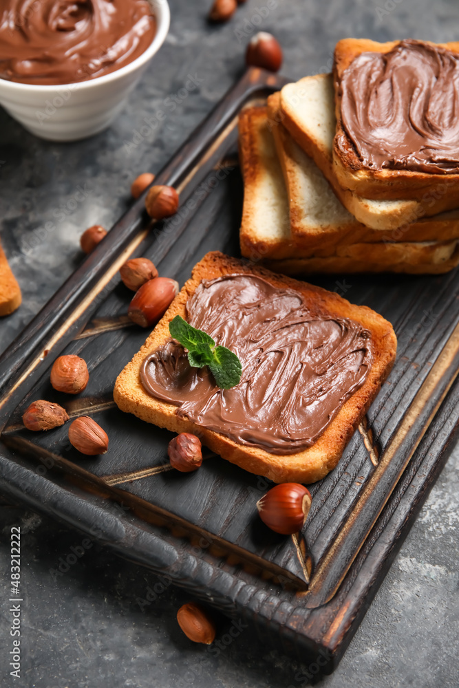 Wooden board of bread with chocolate paste and hazelnuts on black background, closeup