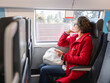 © Konstantin Aksenov - Smiling woman in red duffle coat with on smartphone sits near window in suburban train. Travel by land vehicle.