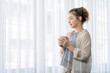 © Jirakul - A woman is sitting in front of the window with a cup in her hand.