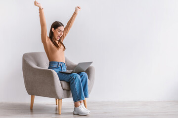 Excited young Caucasian lady sitting in armchair with laptop, lifting hands up, celebrating success against white wall