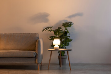 Wall Mural - Comfortable sofa, glowing lamp in evening, potted plant on floor and table in living room interior