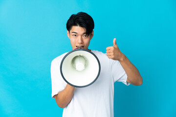 Poster - Young Chinese man isolated on blue background shouting through a megaphone to announce something and with thumb up