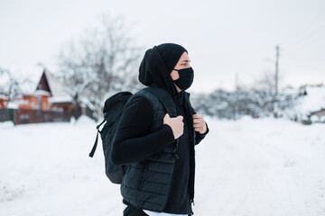 Handsome young fashion man with protective black mask in trendy black clothes with a backpack travels in winter season with snow in town. Coronavirus and style