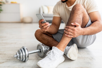 Wall Mural - Closeup of young Arab man sitting on floor with smartphone after dumbbell workout, checking new sports video online