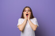 © Red Cristal - Shocked and concerned girl holding hands on face, open mouth and furrow eyebrows startled, hear awful terrible news, standing in blank white t shirt over purple background