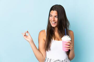 Young brazilian woman with strawberry milkshake isolated on blue background pointing finger to the side and presenting a product