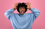 Fototapeta  - Cool African American teen guy making silly gesture with his hands, grimacing and having fun on pink studio background