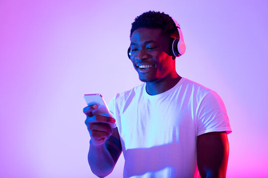 Cheerful black guy in wireless headphones using smartphone in neon light