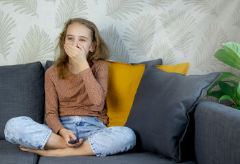 a little girl, sitting on the couch and holding a remote control in her hand while watching TV, covers her mouth with her hand in surprise