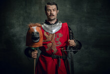 Portrait Of Medieval Warrior Or Knight Wearing Helmet And Armor Riding Toy Horse, Holding Big Sword Isolated Over Dark Background. Comparison Of Eras, History