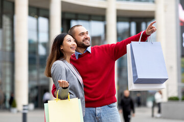 Wall Mural - Positive diverse couple walking along street with paper bags, pointing at new store or advertisement, shopping together