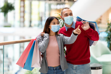 Wall Mural - Portrait of millennial diverse couple with gift bags and face masks window shopping, choosing goods at city mall