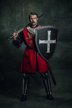 Portrait of medieval warrior or knight with dirty wounded face holding shield and sword isolated over dark background. Comparison of eras, history